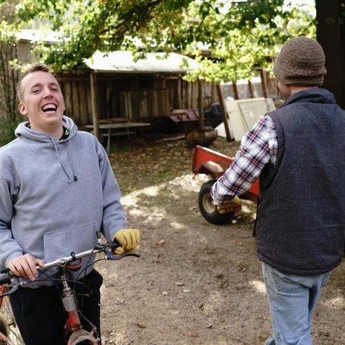 Young man smiling while holding a bicycle outdoors, with another person walking nearby.