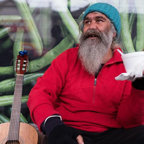 Bearded man in a red jacket sitting with a guitar nearby, holding takeaway food and drink.