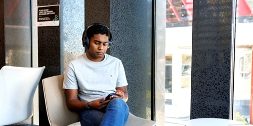 Young man sitting in a chair by a window, wearing headphones and looking at his phone.