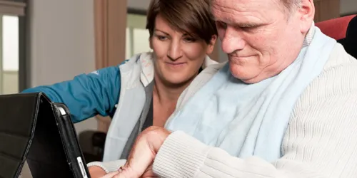 Woman assisting an older man as he uses a tablet, both focused on the screen.