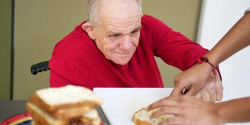 Older man preparing sandwiches with assistance from another person.