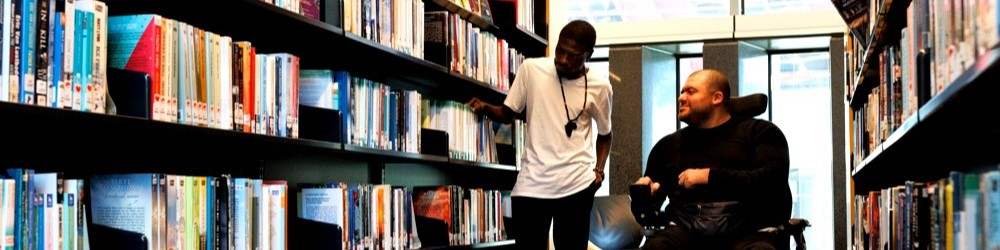 Two men in a library aisle looking at books and talking.