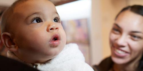 Baby looking upward while a woman smiles at them in the background.