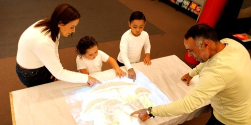 Two adults and two children gathered around a table, pointing at an interactive projection of mushrooms.