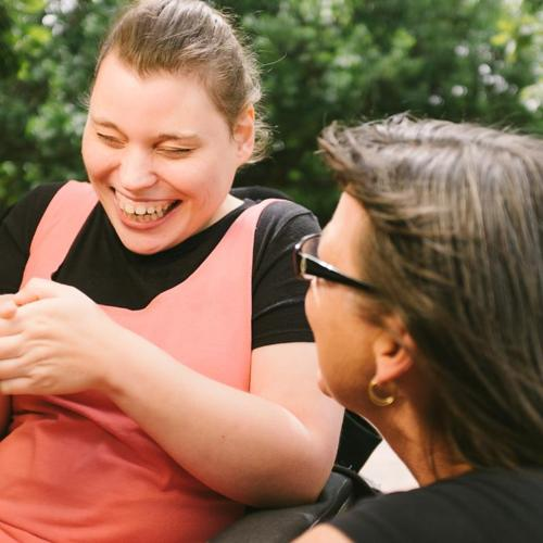 Two women outdoors sharing a laugh, one seated and one leaning in, both smiling warmly.