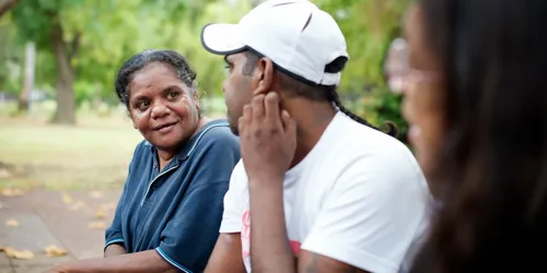 Three people sitting on a park bench talking, one gesturing while listening.
