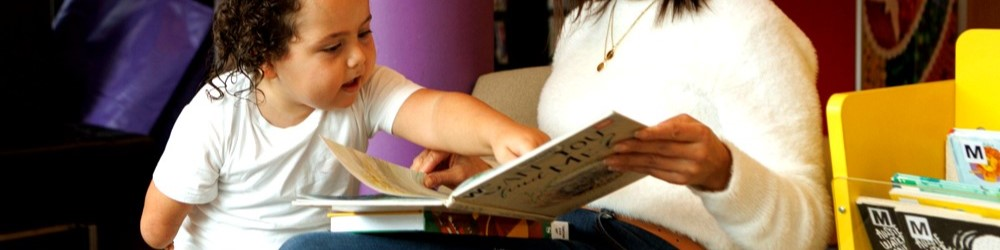 A standing child points to a picture in a book a seated adult is holding in their lap.
