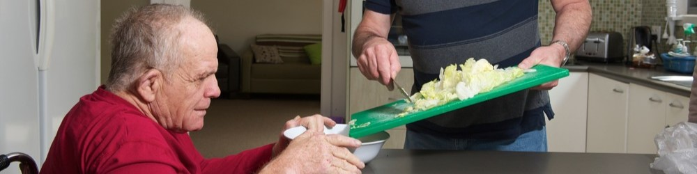 A man preparing food while an older man sits at a table holding a bowl.