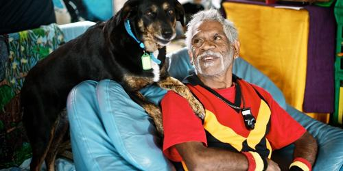 Elderly man sitting on a couch with a dog resting its paws on his shoulder, both looking content.