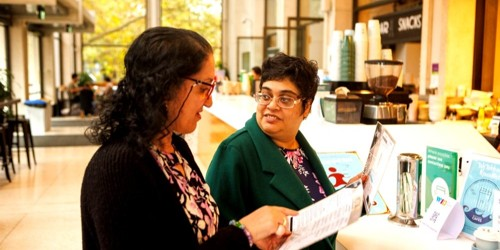 Two women standing at a counter, looking at and discussing printed materials.