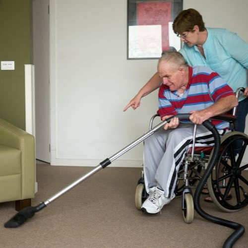 A caregiver assists an older man in a wheelchair as he uses a vacuum cleaner.