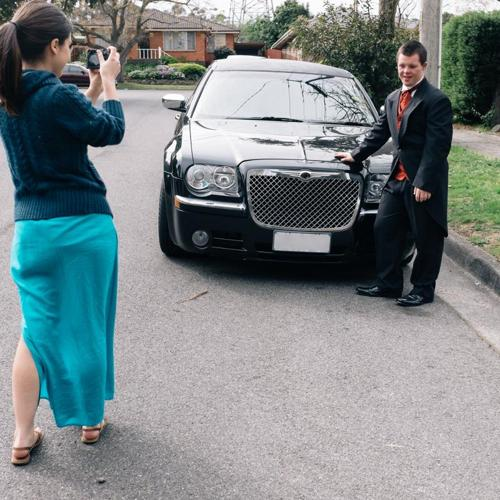 Person in formal attire standing beside a car while another person takes their photo on a street.