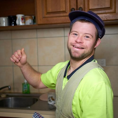 Smiling man in a kitchen wearing a green shirt and cap, mid-gesture.
