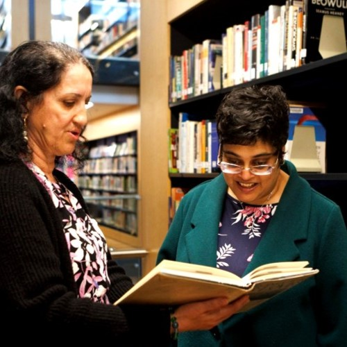 Two women standing in a library, smiling as they read a book together.