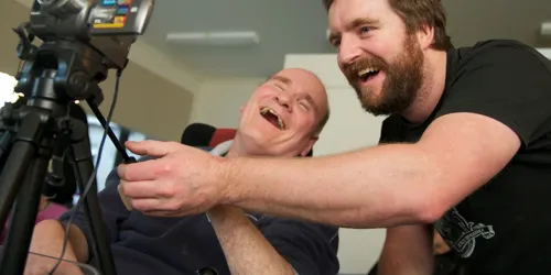 Two men indoors laughing while looking at a camera on a tripod.