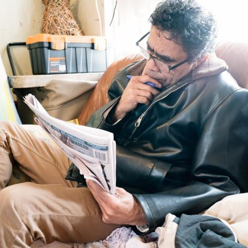 Man sitting on a couch reading a newspaper while holding a pen, appearing focused.