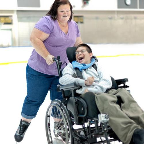 Woman assisting a person in a wheelchair on an ice rink, both smiling.
